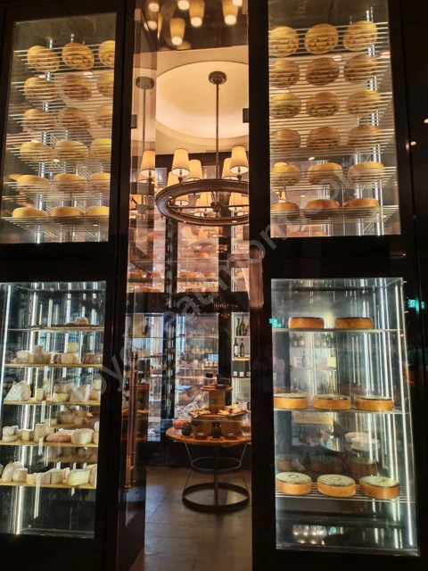 Glass-fronted cheese room with illuminated shelves and a central tasting table.