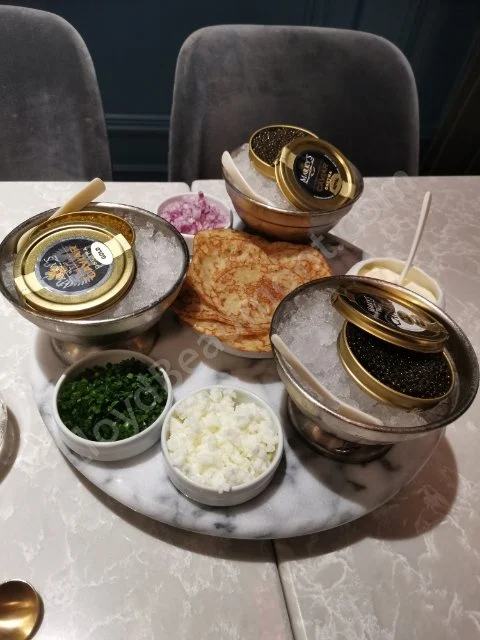 Caviar tins on ice with blinis and traditional accompaniments on a marble serving tray.