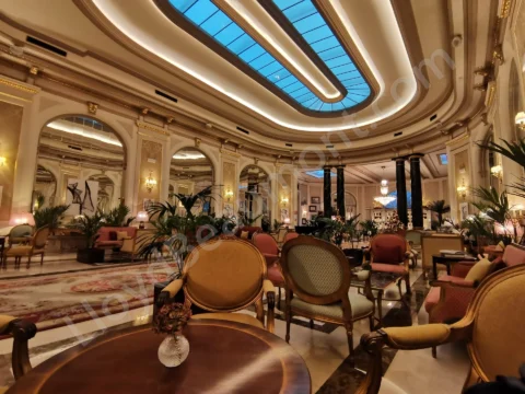 Grand hotel lounge with pastel chairs, marble floors, chandeliers, and a blue-lit skylight.