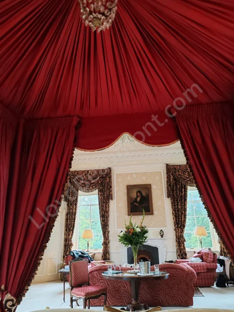Rich red canopy curtains frame an elegant suite with a fireplace, flowers, and upholstered seating.
