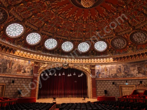Ornate concert hall with a richly decorated domed ceiling, red seats, and a curtained stage.