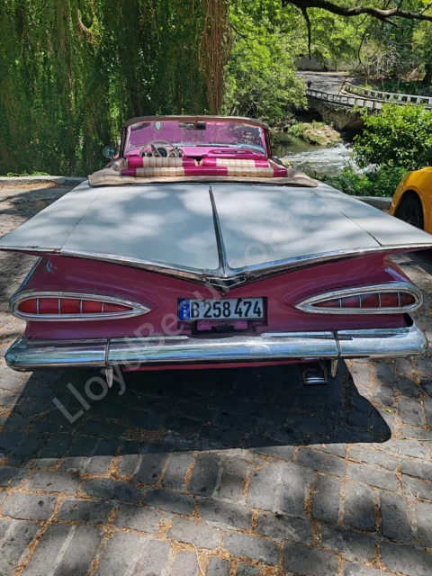 Pink vintage convertible parked beside a leafy stream and stone bridge.