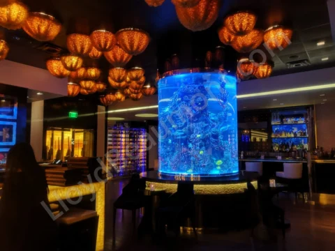Blue-lit cylindrical aquarium at the center of a warmly illuminated cocktail lounge.