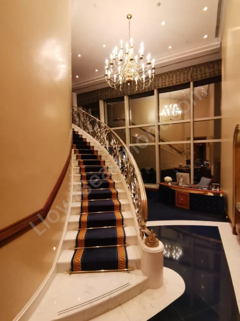 Curving marble staircase with a blue runner, gilded railing, and chandelier inside a hotel suite.