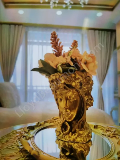 Gold sculptural vase with flowers on a mirrored tray, with a softly lit bedroom beyond.