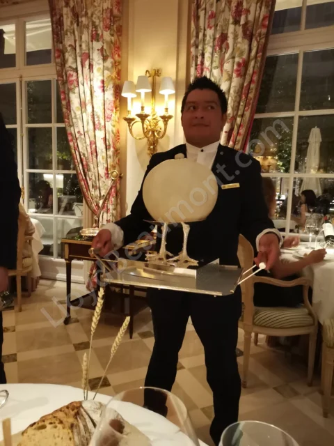 Tableside cheese presentation in an elegant dining room with floral curtains and warm lighting.
