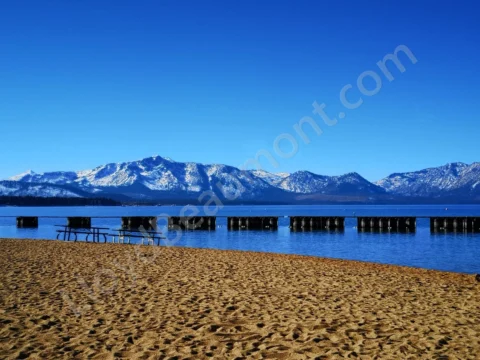 Sandy lakeshore and wooden piers facing snow-covered mountains beneath a clear blue sky.