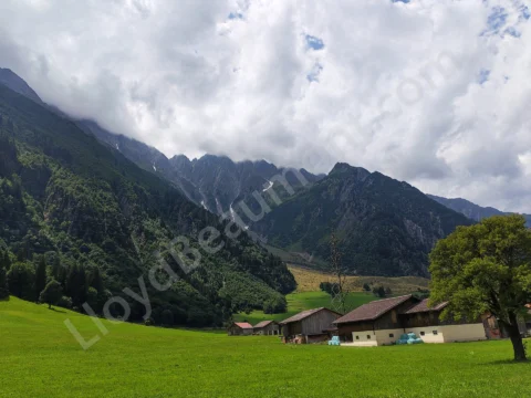 Lush mountain meadow with farm buildings below rugged, cloud-covered alpine peaks.