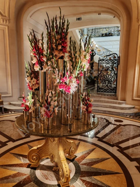 Tall pink-and-white floral arrangements in gold vases beside an ornate hotel staircase.